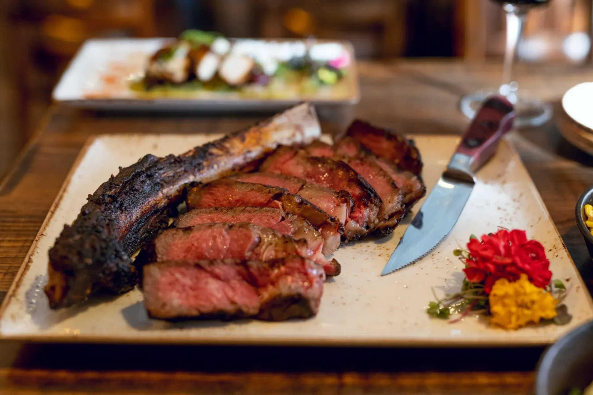 A sliced bone-in steak on a plate with a knife, flowers, and a glass of red wine